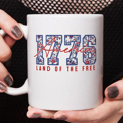 White mug with '1776 America Land of the Free' design held by a person.