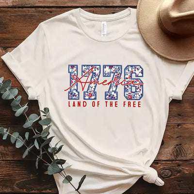 White t-shirt with '1776 America Land of the Free' design on a wooden surface with a hat and plant.