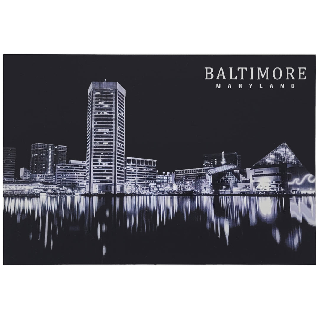 A black and white postcard of the Baltimore, Maryland harbor skyline at night, featuring illuminated buildings and their reflections on the water, with the text &