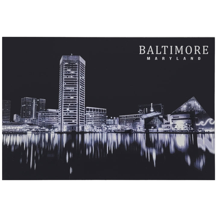 A black and white postcard of the Baltimore, Maryland harbor skyline at night, featuring illuminated buildings and their reflections on the water, with the text &