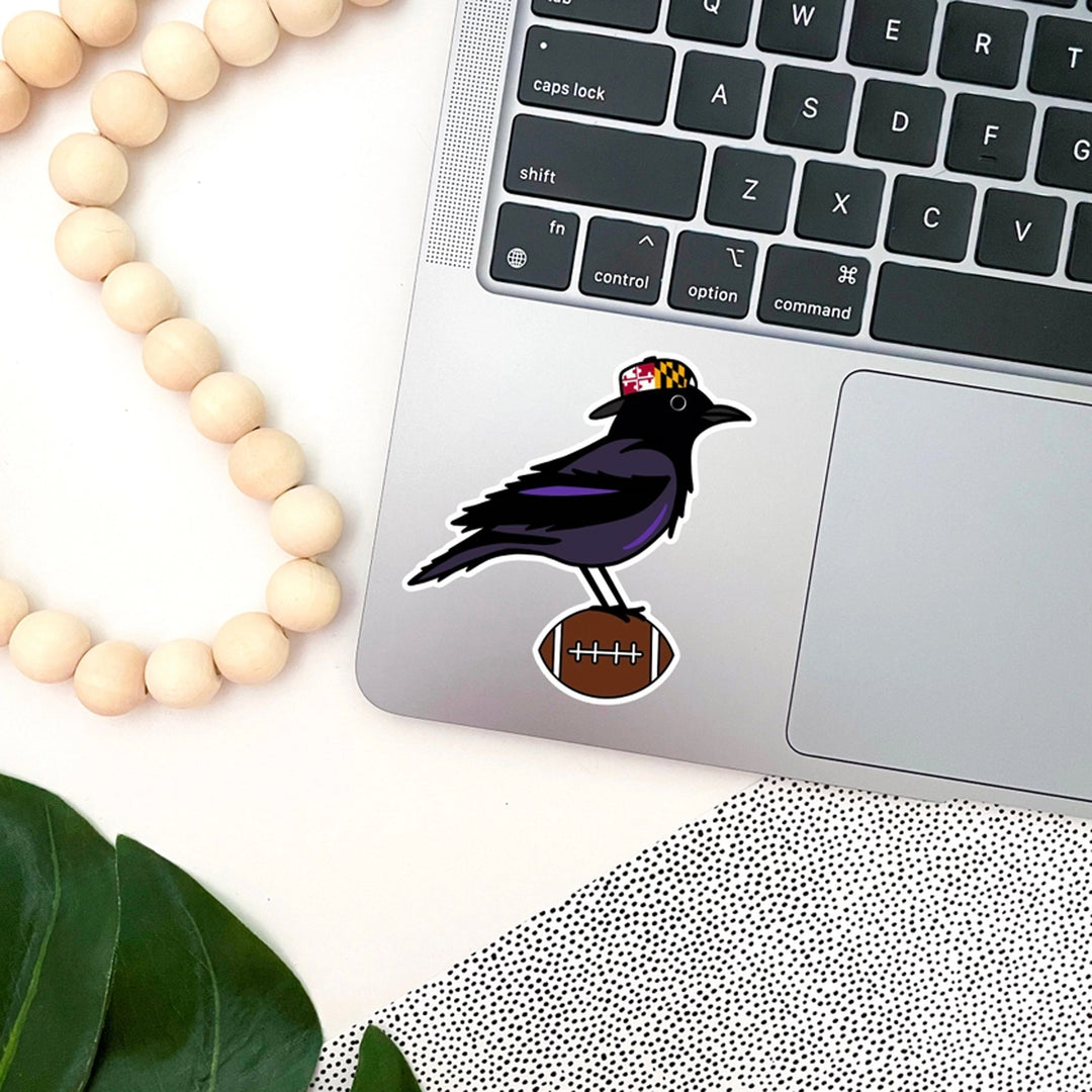 Laptop with a sticker of a black raven bird holding a football, surrounded by wooden beads and a plant leaf.