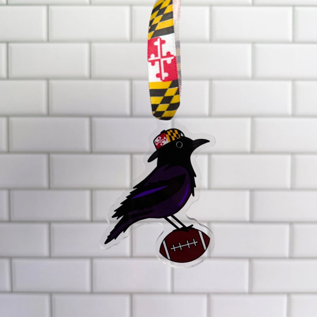 A Maryland Ravens themed ornament featuring a black and purple raven wearing a Maryland flag cap and standing on a football, hanging from a Maryland flag ribbon against a white subway tile wall.