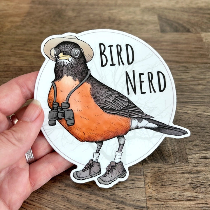 A die-cut sticker featuring a cartoon robin wearing a beige hat, black binoculars around its neck, white socks, and hiking boots, with the words “BIRD NERD” in black text on a white circular background.