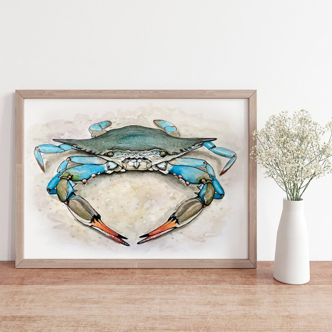 A framed art print of a blue crab, styled on a light wood surface next to a white ceramic vase with dried baby&