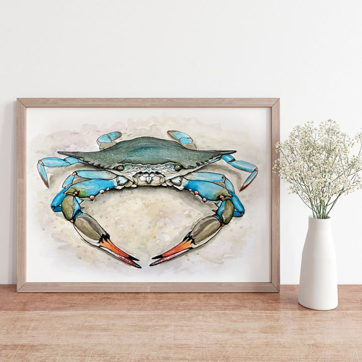 A framed art print of a blue crab, styled on a light wood surface next to a white ceramic vase with dried baby&