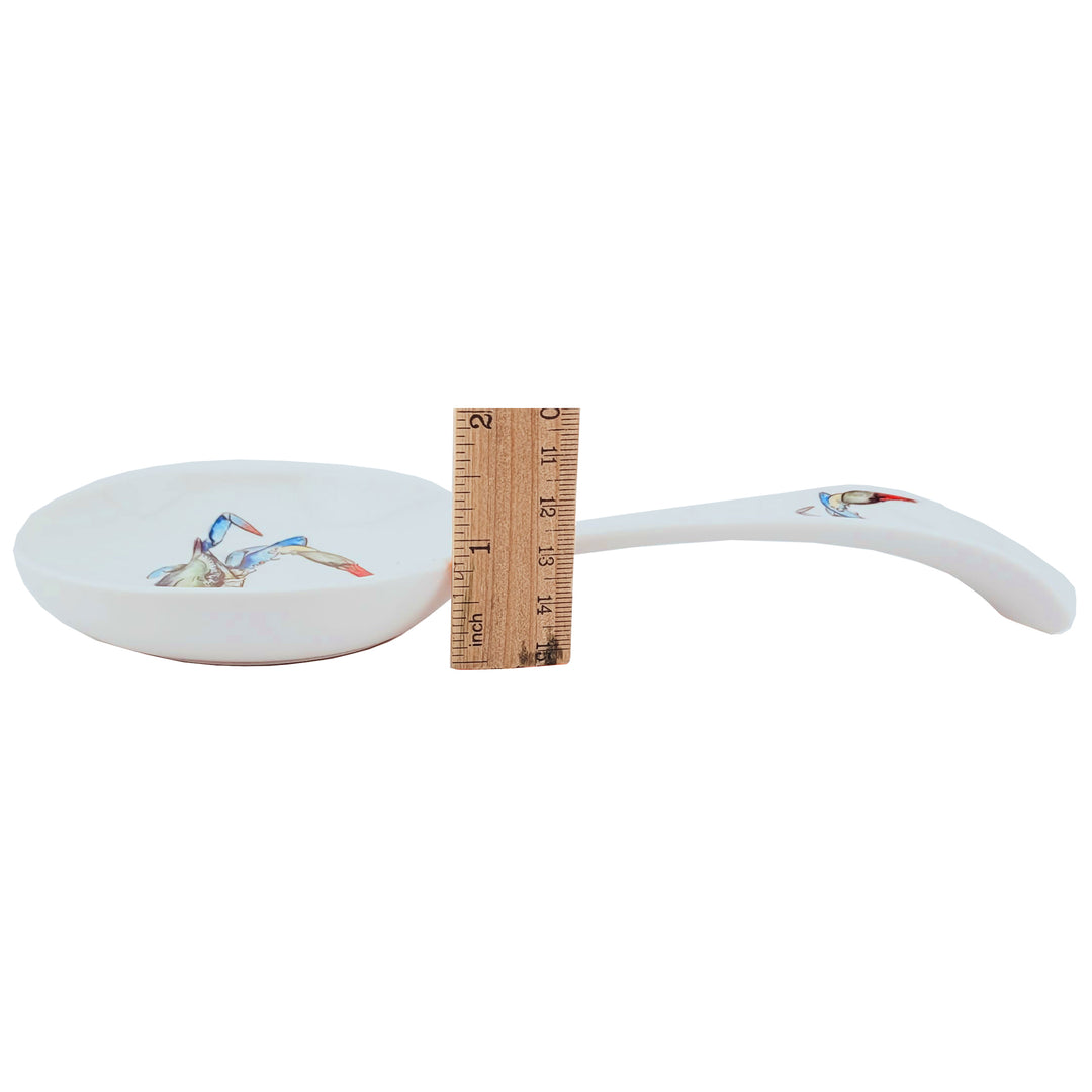 A white ceramic spoon rest with a blue crab illustration on the bowl and handle. shown next to a wooden ruler for scale.