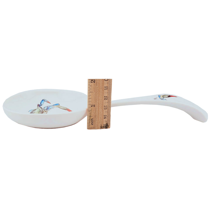 A white ceramic spoon rest with a blue crab illustration on the bowl and handle. shown next to a wooden ruler for scale.