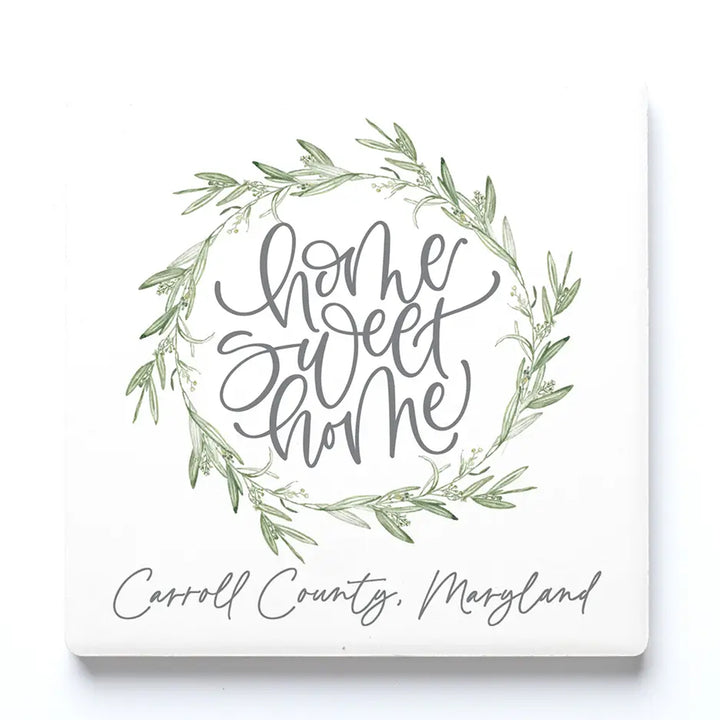 A square ceramic drink coaster with a green leaf wreath and gray script reading “home sweet home,” with “Carroll County, Maryland” written below in gray script.