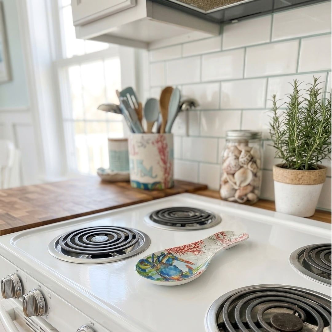 A white ceramic electric stovetop with black coil burners in a bright kitchen setting, featuring a decorative blue crab coastal design spoon rest.