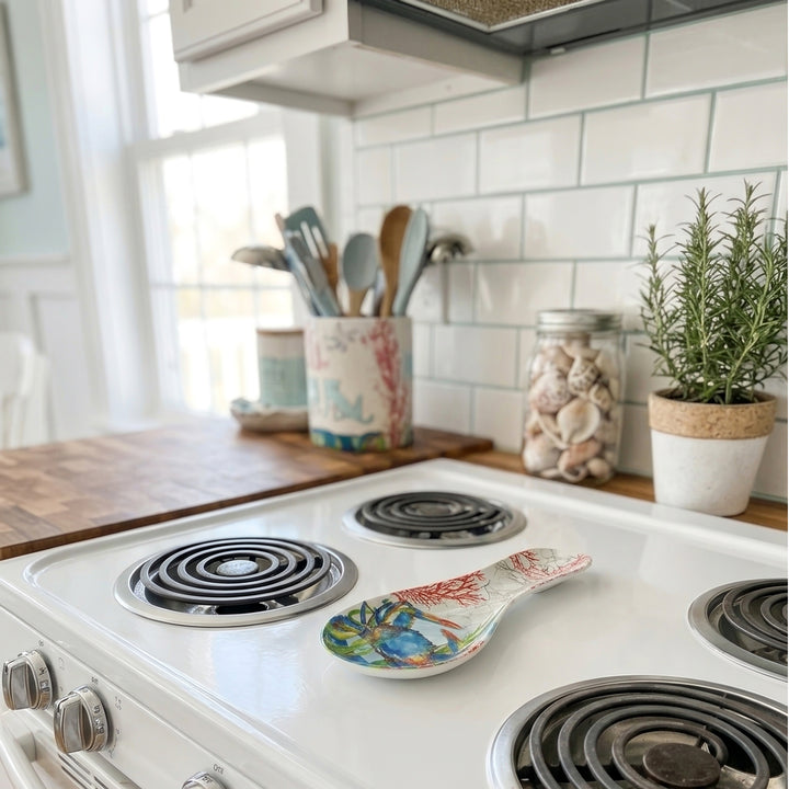A white ceramic electric stovetop with black coil burners in a bright kitchen setting, featuring a decorative blue crab coastal design spoon rest.