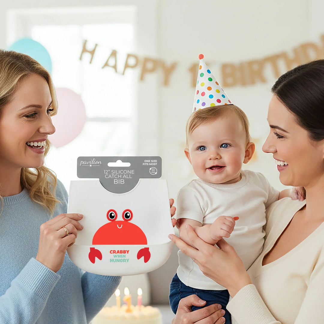 Two women holding a baby and a silicone baby bib with a crab design in a celebratory setting.