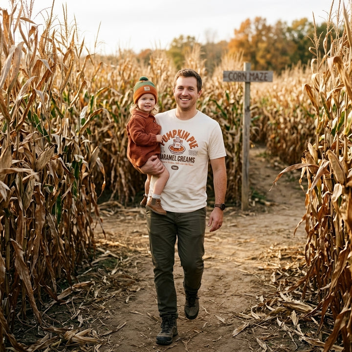 A man walking in a corn maze path while holding a toddler. The man is wearing a cream graphic t-shirt that reads &
