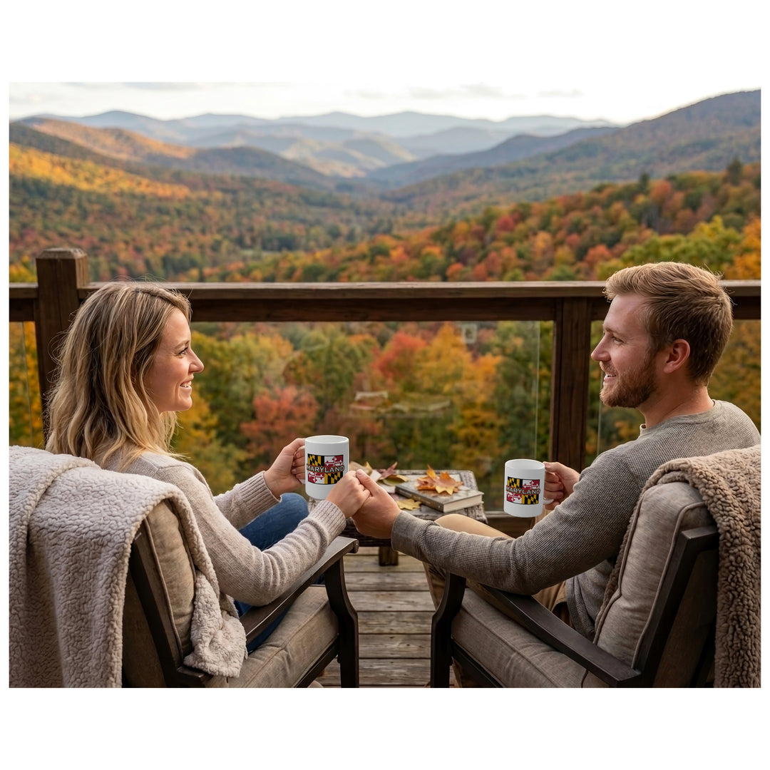 A man and a woman sit on outdoor chairs set on a balcony, holding Maryland flag mugs, and overlooking  a colorful mountain valley in the fall.