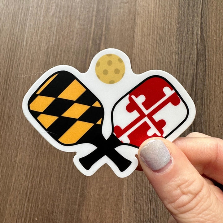 A die-cut sticker featuring crossed pickleball paddles with the Maryland state flag design on one paddle and a red cross on the other, with a small yellow pickleball above them on a white background.