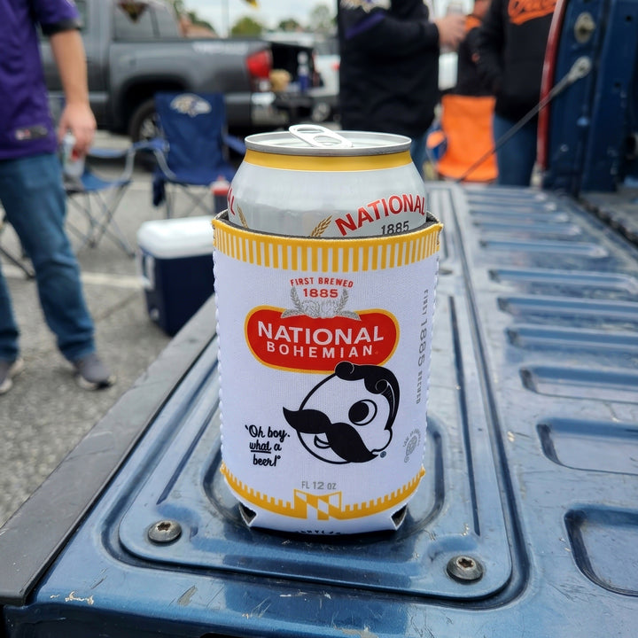 A Natty Boh Can Coolie sitting on the tailgate of a blue pickup truck at an outdoor event.