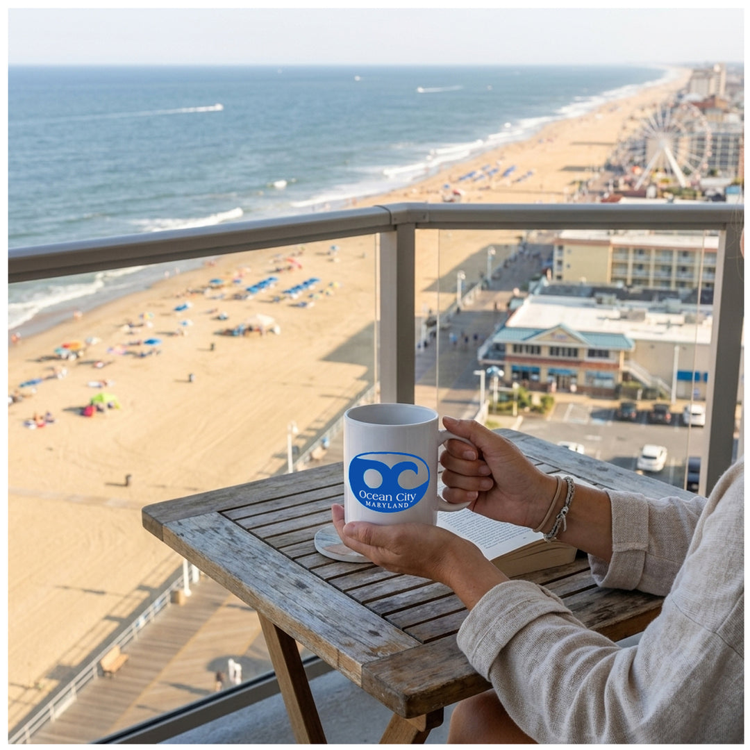 A person sitting on a balcony overlooking the Ocean City Maryland beach holds a white ceramic mug with a blue Ocean City Maryland wave logo.