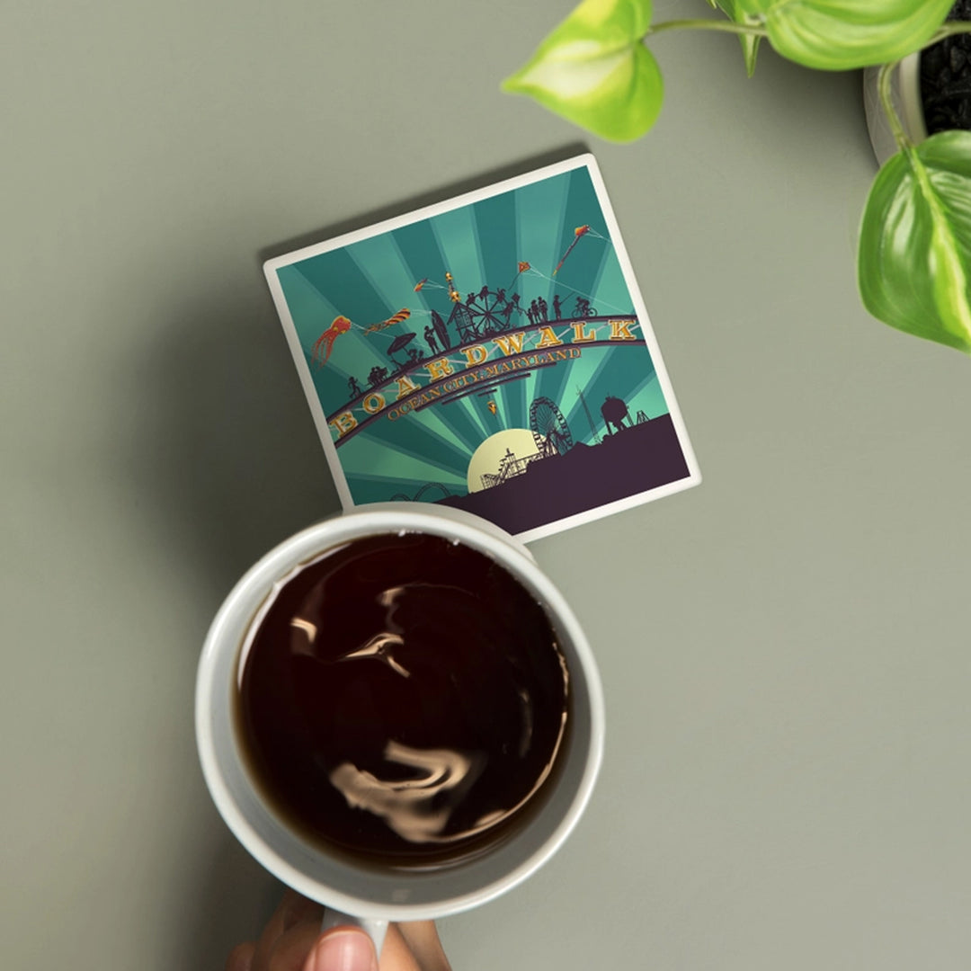 A ceramic drink coaster featuring a stylized Boardwalk, Ocean City, Maryland scene with silhouettes of people, kites, amusement park rides, and sunburst rays.