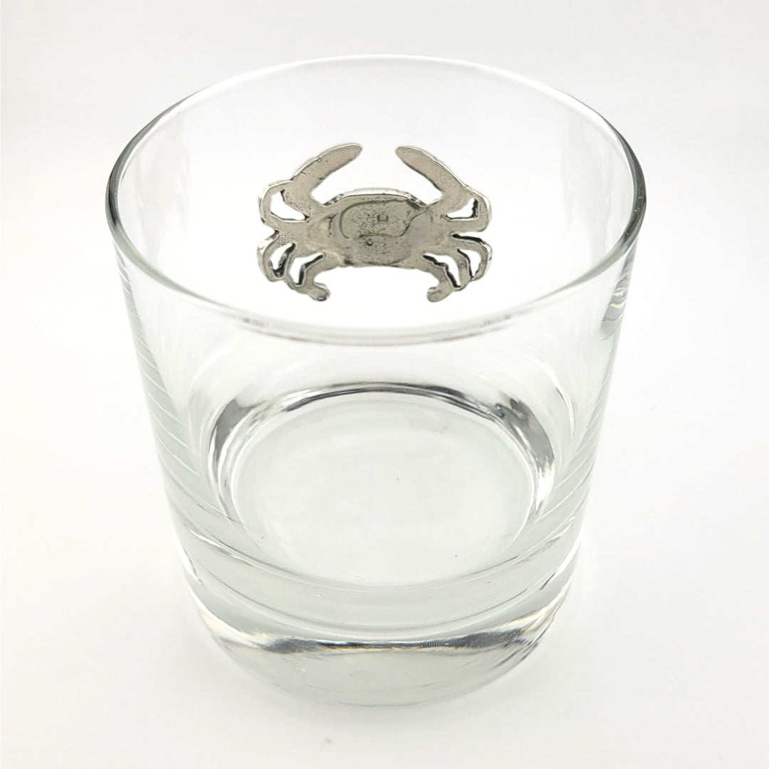 A clear glass rocks tumbler with a pewter crab emblem centered on the upper rim, photographed on a clean white background.