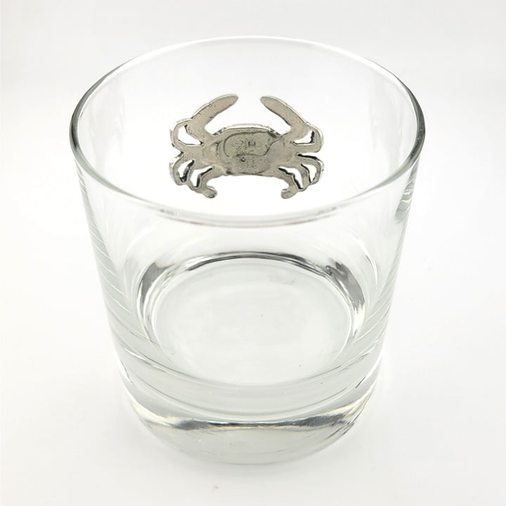 A clear glass rocks tumbler with a pewter crab emblem centered on the upper rim, photographed on a clean white background.
