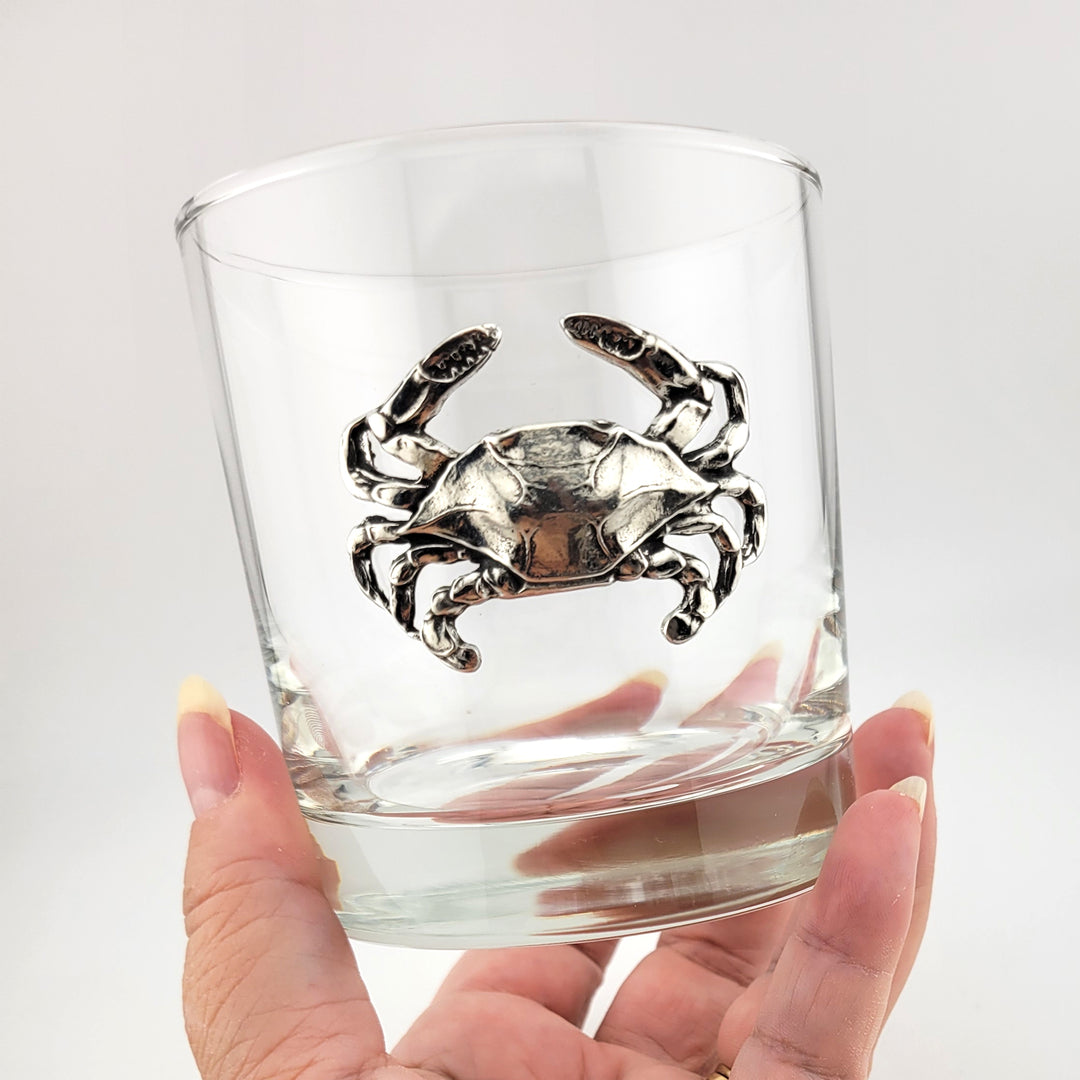 A hand holding a clear rocks glass featuring a detailed silver crab motif on the front, set against a plain light gray background.