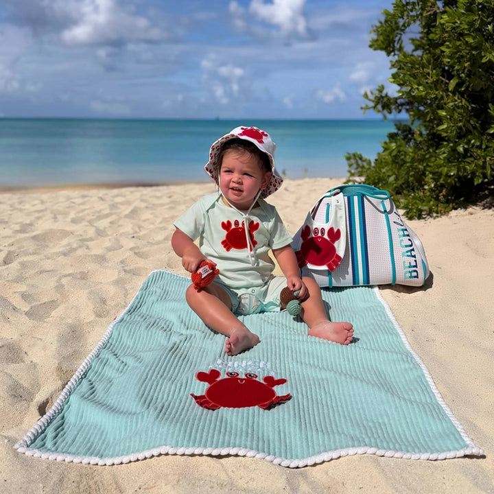 Child sitting on a Pinch Perfect Blanket with crab design, wearing a hat and holding a toy, with a beach bag in the background.