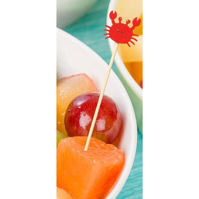 Fruit skewer with a crab-shaped toothpick in a bowl of fruit on a turquoise surface