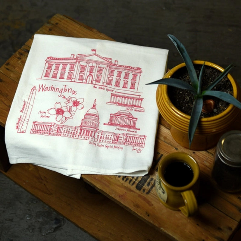 White towel with red graphics of Washington DC landmarks on a wooden table with a mug and plant.